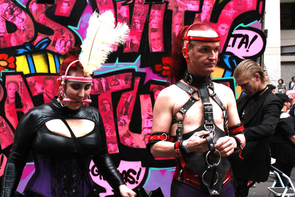 Couple wearing advanced bondage gear with leather harnesses and restraints at a fetish gathering.