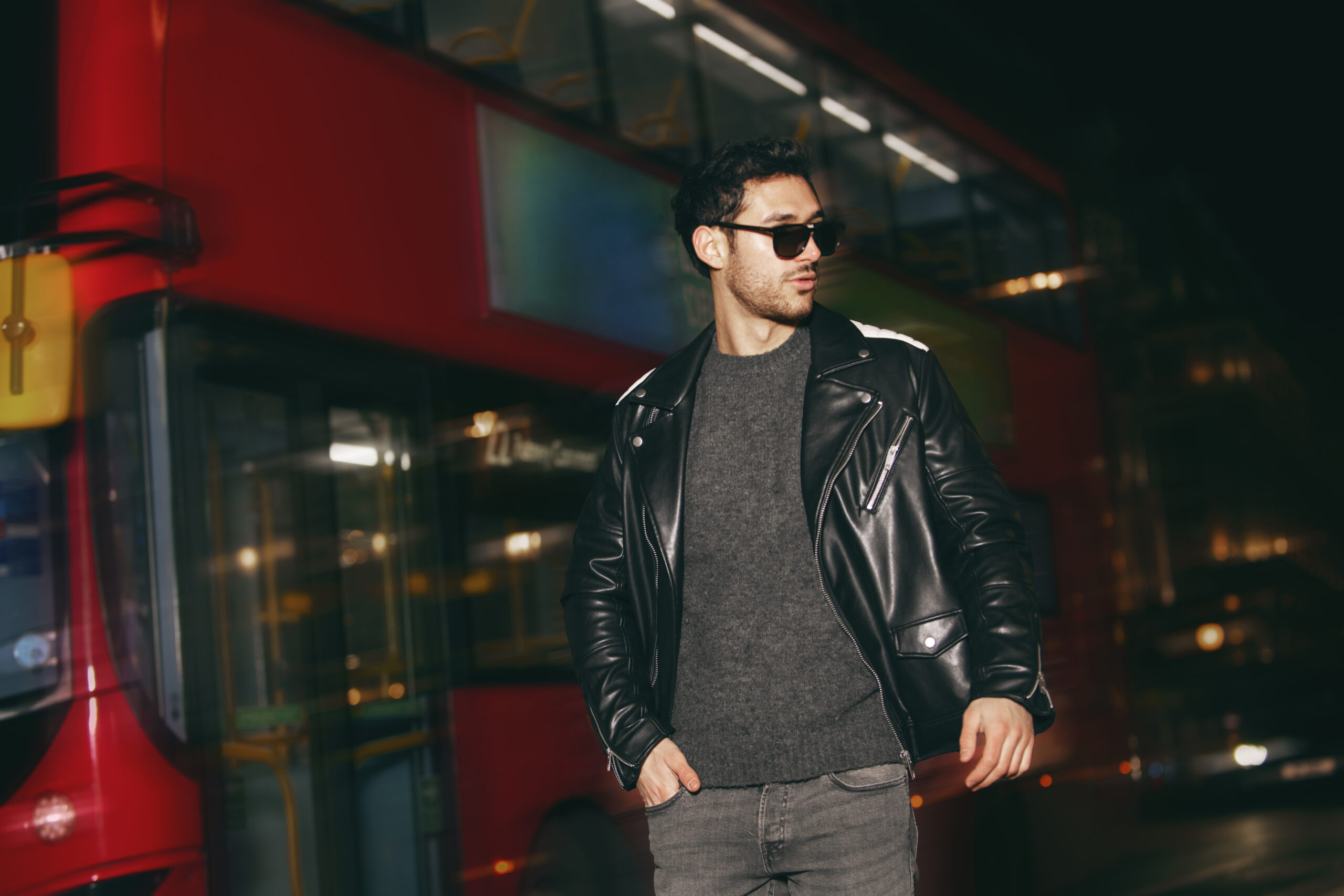 "Man wearing a black leather jacket and sunglasses standing in front of a red double-decker bus at night."